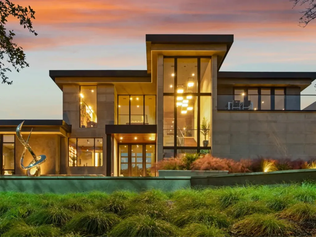 Modern two-storey home with floor-to-ceiling windows glowing at dusk behind a landscaped garden